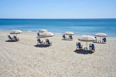Sandy beach with organized seating areas featuring white umbrellas and blue lounge chairs along calm blue ocean waters