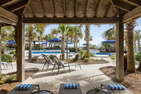 Wooden pergola structure overlooking resort pool area with palm trees, blue umbrellas, and lounge chairs