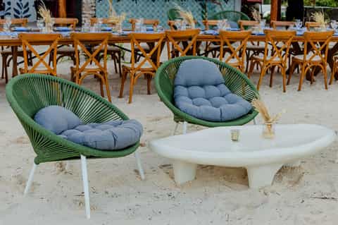 Beach lounge area with green woven pod chairs, white round table, and long dining setup beyond