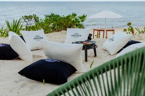Beach lounge area with white and black pillow cushions labeled 'Volcan', umbrella, and ocean view