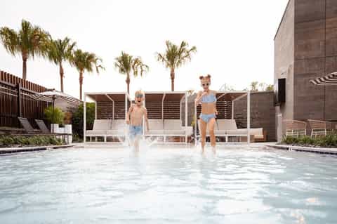 Children running and splashing in shallow resort pool with palm trees and modern structures