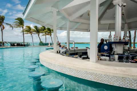 Modern pool bar under white pergola with turquoise water, palm trees, bartender, and tropical waterfront view