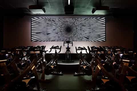 Modern fitness studio with rows of stationary bikes facing large hypnotic light display screen