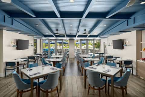 Modern restaurant dining room with blue chairs, white tables, lattice ceiling, and ocean views through glass doors