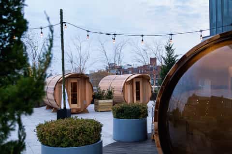 Rooftop sauna pods with wooden barrels, string lights, greenery, and city skyline backdrop