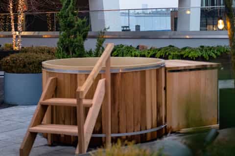 Wooden hot tub on terrace with steps, planters, illuminated landscaping, and evening city views