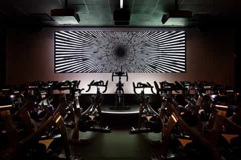 Fitness studio with rows of stationary bikes and large geometric light display wall