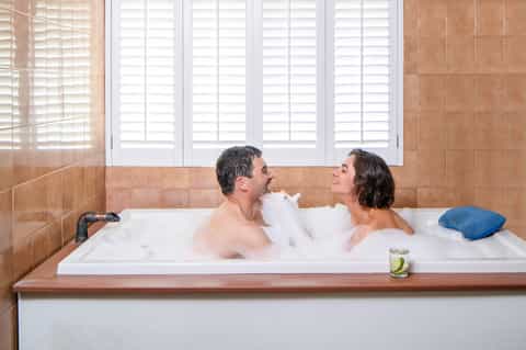 Couple relaxing in a large white bathtub with white shuttered windows and terracotta tile walls