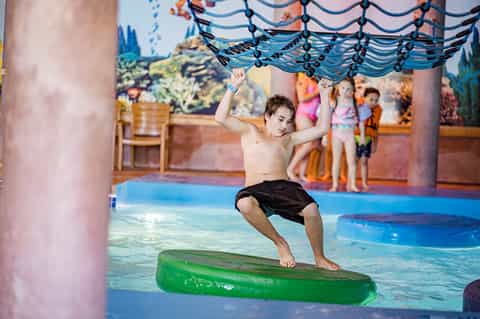 Child on green floating device in indoor pool facility with rope obstacle course and murals