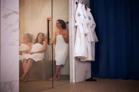 Women enjoying spa steam room with marble walls and monogrammed white robes hanging nearby