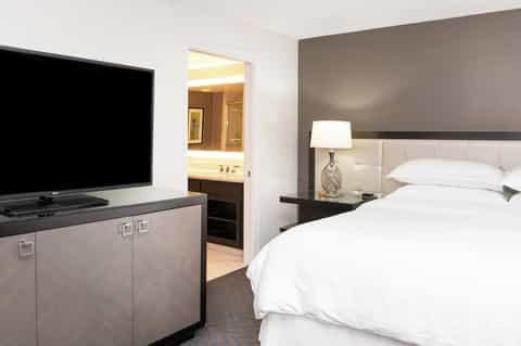 Hotel bedroom with TV, white bedding, gray headboard, and open bathroom door