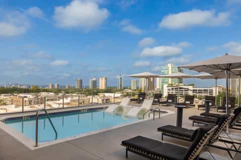 Rooftop pool overlooking city skyline with loungers, umbrellas, and blue-tinted water on sunny day