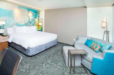 Hotel room with king bed, ocean-inspired headboard art, sofa, and turquoise accent pillows