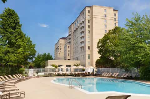 Multi-story hotel with outdoor swimming pool, lounge chairs, and mature trees on sunny day