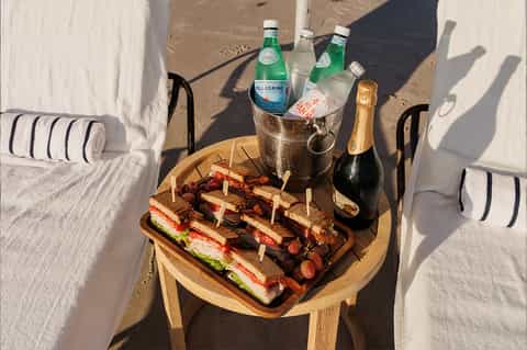 Beach loungers with champagne, sparkling water, and gourmet sandwiches on wooden table