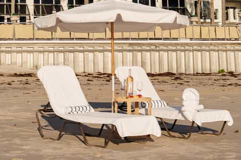 Beach lounge setup with two white chairs, umbrella, small table with wine bottle and glasses