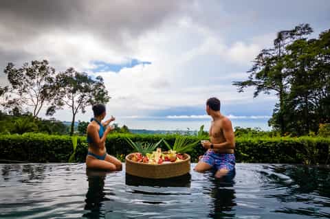 Two people in infinity pool sharing floating fruit basket with lush tropical landscape background
