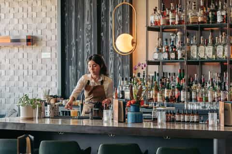 Contemporary bar interior with bartender behind counter, extensive bottle display, and modern lighting