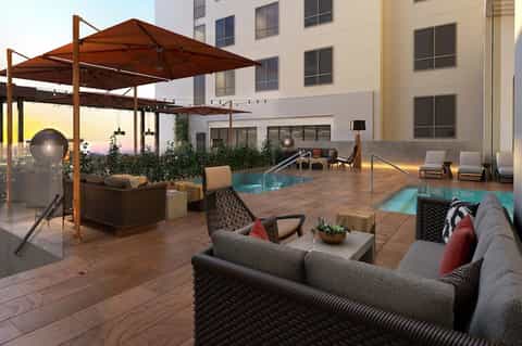 Rooftop lounge at dusk with pools, seating areas, pergola, and city building backdrop