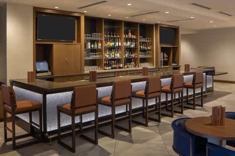 Hotel lounge with long wooden bar, leather chairs, backlit liquor bottles, and flat-screen TV