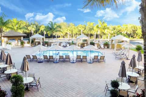 Large resort pool surrounded by white cabanas, palm trees, and gray umbrellas in sunny courtyard