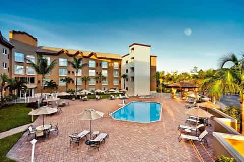 Resort exterior with multi-story building, pool area with loungers, umbrellas, and tropical landscaping