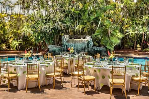 Elegant poolside banquet setup with white linens, gold chairs, and tropical waterfall backdrop