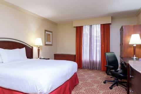 Traditional hotel room with burgundy accents, wooden furniture, queen bed, and red drapes framing windows