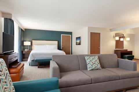 Hotel suite with teal accent wall, queen bed, sofa, and flat-screen TV in neutral tones