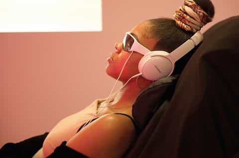 Person wearing white headphones and pink sunglasses relaxing against pink background wall