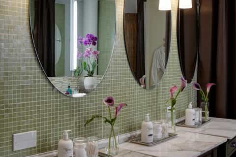 Luxury spa bathroom with dual oval mirrors, green tile backsplash, purple orchids, and marble countertop
