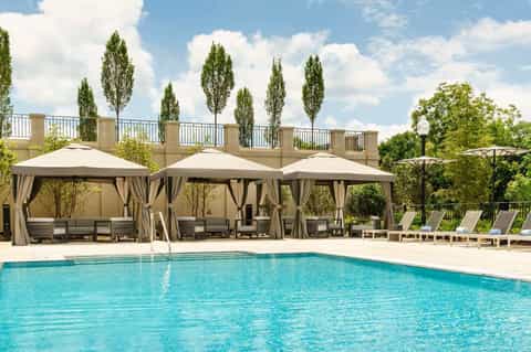 Resort pool with beige cabanas featuring curtains, tall cypress trees, and landscaped grounds