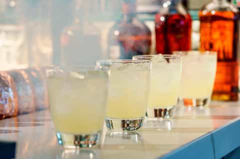 Row of clear glasses with pale yellow cocktails on a bar with blurred bottles in background