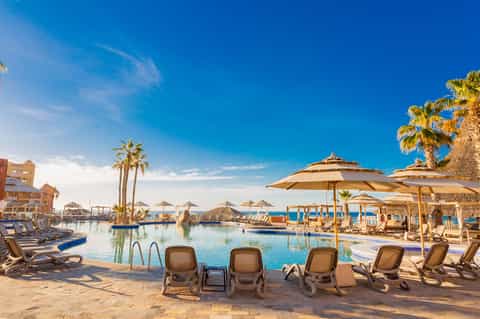 Tropical resort pool complex with lounge chairs, umbrellas, palm trees, and ocean views at day