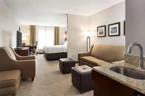 Spacious hotel suite featuring bed, sitting area with tan leather sofa, kitchenette with granite countertop, and flat-screen TV.