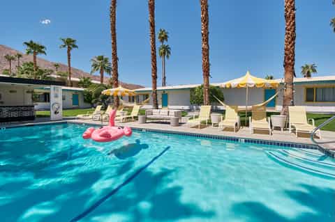 Retro-style pool with pink floats, palm trees, yellow umbrellas, and mid-century modern cabanas