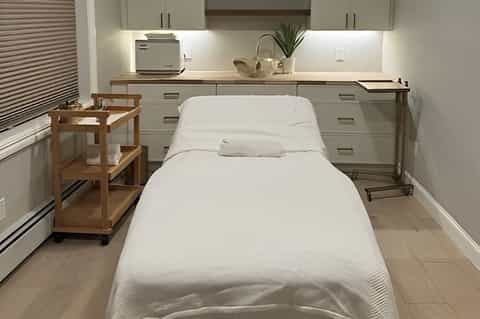 Minimalist spa treatment room with massage bed, vanity, wooden shelving, and clean white decor