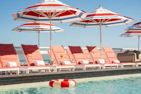 Resort pool deck with red and white striped umbrellas, lounge chairs, and ocean backdrop