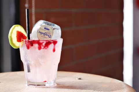 Cocktail with lime wedge, red syrup drizzle, and ice in clear glass on wooden table