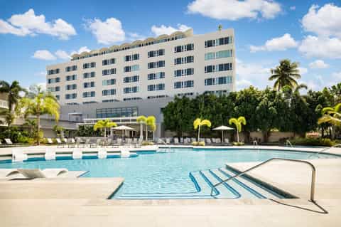 Large modern hotel with multi-story white facade, beach-style pool with loungers and palm landscaping