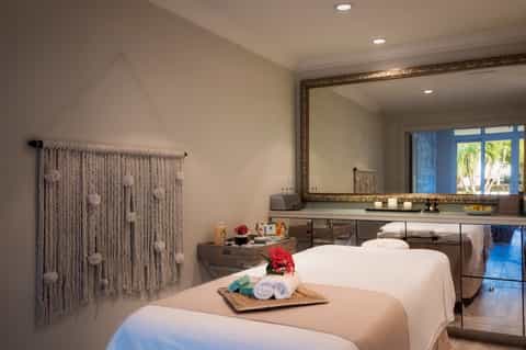 Spa treatment room with two massage tables, framed mirror, and macramé wall hanging