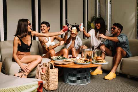 Group of five friends celebrating on outdoor sectional with food, drinks, and fire pit table