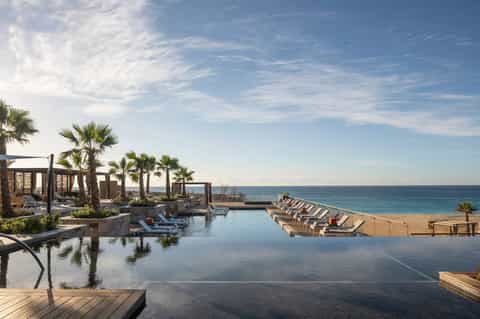 Beachfront infinity pool overlooking ocean, palm trees, cabanas, and sandy beach with loungers