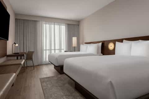 Modern hotel room with dual beds, wood headboard, floor-to-ceiling curtains, and desk area