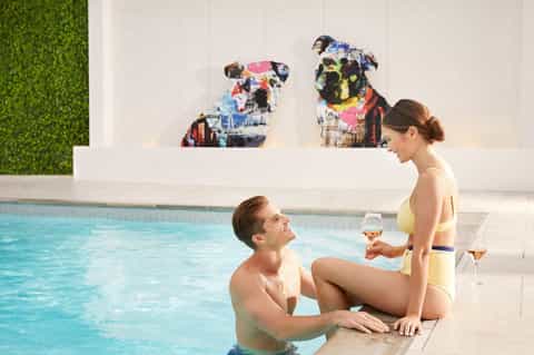 Indoor pool with modern wall art, man in water talking to woman at poolside holding wine glass
