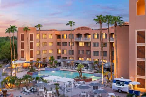 Resort courtyard at dusk with illuminated buildings, pool, palm trees, and outdoor dining area