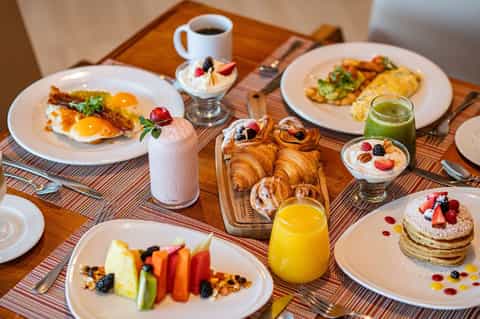Hotel breakfast spread with eggs, croissants, pancakes, fresh fruit, juice, and coffee on wooden table
