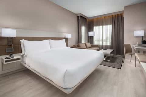Modern hotel bedroom with king bed, seating area, and floor-to-ceiling windows