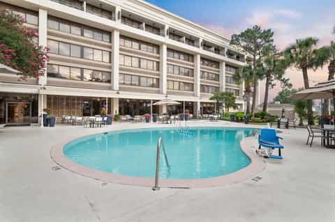 Modern resort pool with turquoise water surrounded by white concrete, lounge chairs, multi-story building facade, and palm trees