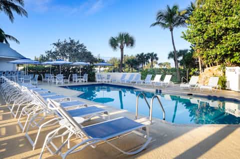 Resort pool area with white loungers, blue umbrellas, palm trees, and manicured tropical grounds